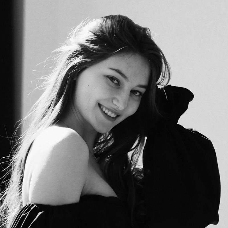 Black-and-white portrait of a young woman with long hair, smiling in a casual black top, captured in natural light.